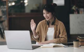 Asian woman wearing headphones on her neck using laptop and greeting friends on video call at home photo