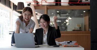 Two young Asian businesswomen show joyful expression of success at work smiling happily with a laptop computer in a modern office. photo