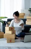 una joven y sonriente propietaria de una empresa asiática prepara una caja de paquetes y revisa de pie los pedidos de productos en línea para entregarlos al cliente en una computadora portátil. concepto de compras en línea. foto