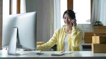 un retrato de una mujer asiática, empleada de comercio electrónico sentada en la oficina llena de paquetes en la mesa usando una computadora portátil y un teléfono inteligente, para pymes, comercio electrónico, tecnología y negocios de entrega. foto