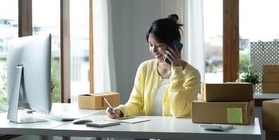 A portrait of Asian woman, e-commerce employee sitting in the office full of packages on the table using a laptop and smartphone, for SME business, e-commerce, technology and delivery business. photo