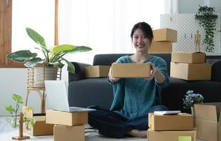 Happy young asian woman startup small business freelance holding parcel box and computer laptop and sitting on floor, Online marketing packing box delivery concept photo