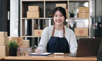 A portrait of a young Asian woman e-commerce employee sitting in the office full of packages in the background write note of orders and a calculator, for SME business e-commerce and delivery business.