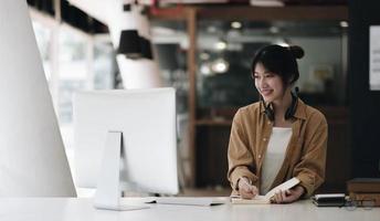 Asian woman wearing headphones using laptop and taking notes on homework that her teacher teaches at home photo