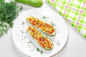 Top view of baked zucchini halves with vegetables, couscous and greens on a white wooden background with fresh zucchini and dill photo