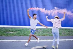 Young couple riding on skateboards with color smoke bomb, boy and girl in casual clothes having good time, pink and blue colors photo