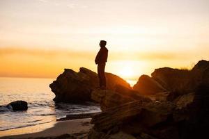la silueta de un viajero masculino se para cerca del acantilado y observa la hermosa puesta de sol en el mar foto