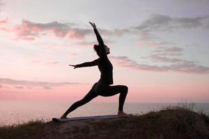 Fit woman doing yoga stretching exercise outdoor in beautiful mountains landscape. Female on the rock with sea and sunrise or sunset background training asans. Silhouette of woman in yoga poses photo