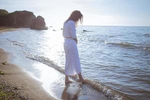 mujer joven caminando en la playa de la mañana en un hermoso vestido blanco. mujer en forma pasando un buen rato durante el amanecer. foto