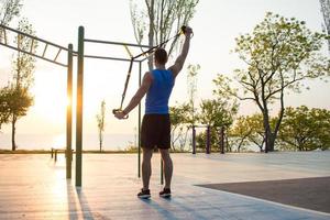 workout with suspension straps In the outdoor gym, strong man training early in morning on the park, sunrise or sunset in the sea background photo