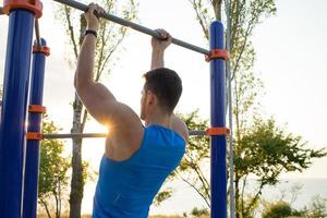 Muscular man doing pull-ups on horizontal bar, training of strongman on outdoor park gym in the morning.