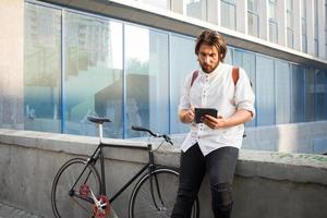 Young bearded man with leather backpack walking with black bicycle photo