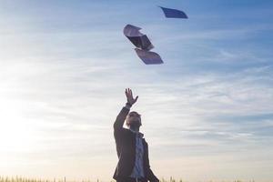 Portrait of young businessman throwing sheets of paper into the air, sunset in summer fields background photo