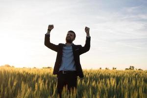 concepto de ganador, joven empresario feliz saltando con las manos levantadas en los campos, plantas de trigo de verano foto