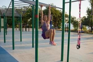 Beautiful fit woman in pink and purple sportwear training on outdoor gym in morning, exercises with suspension straps in park photo