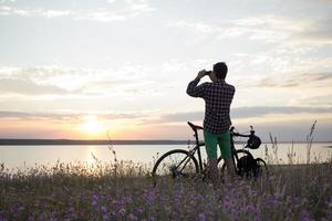 silueta de un hombre con bicicleta de carretera de gira mirando y haciendo una foto de la puesta de sol en el lago en el teléfono celular