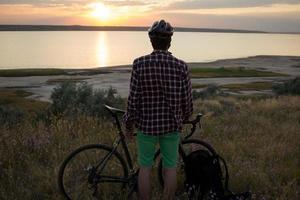 silueta de un hombre con bicicleta de carretera de gira mirando y haciendo una foto de la puesta de sol en el lago en el teléfono celular