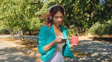 mujer joven con traje colorido y cabello rosado bebe café y escucha música en el parque foto