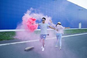 Young couple riding on skateboards with color smoke bomb, boy and girl in casual clothes having good time, pink and blue colors