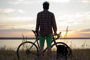 silueta de un hombre con bicicleta de carretera de gira mirando y haciendo una foto de la puesta de sol en el lago en el teléfono celular