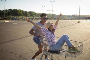 Happy young couple riding on trolley on empty mall parking , hipster friend have good time during shopping, couple in love riding on shopping cart photo