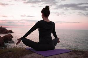 Fit woman doing yoga stretching exercise outdoor in beautiful mountains landscape. Female on the rock with sea and sunrise or sunset background training asans. Silhouette of woman in yoga poses photo