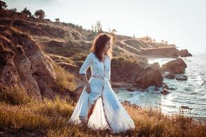 young woman walking on the morning beach in beautiful white dress. Fit female having good time during turing the sunrise. photo