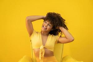 feliz risa mujer africana americana con su pelo rizado sobre fondo amarillo. mujer rizada riendo en suéter tocando su cabello y mirando a la cámara.