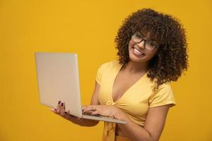 Portrait of a smiling young curly hair mixed race girl holding laptop computer isolated over yellow background. photo