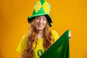 partidario de brasil. hincha brasileña pelirroja celebrando el fútbol, partido de fútbol con fondo amarillo. colores de brasil. con camiseta, bandera y gorra de abanico. foto