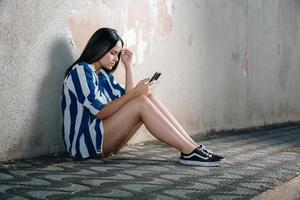 Single sad teen holding a mobile phone lamenting sitting on the sidewalk. Crying depressed teen girl holds phone sitting on the sidewalk. photo