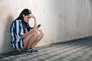 Single sad teen holding a mobile phone lamenting sitting on the sidewalk. Crying depressed teen girl holds phone sitting on the sidewalk. photo