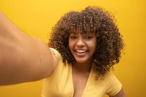 Close up of excited young african american girl in fur sweater posing isolated on yellow orange background in studio. People lifestyle concept. Mock up copy space. Doing selfie shot on mobile phone. photo