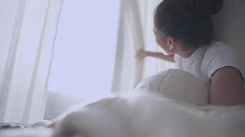 ung asiatisk kvinna vaknar och öppnar gardinerna för att njuta av varmt morgonsolljus, öppna fönstergardiner och titta ut genom fönstret, ny början börja ny dag, solljus blossar upp genom fönstret video