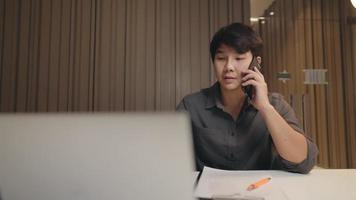 Asiatisk affärsman sitter vid skrivbordet och kommunicerar via telefon medan han stirrar läser på datorn på kontorsrummet sent på natten, övertidsarbete. trött ensam entreprenör man med ögon trötthet video