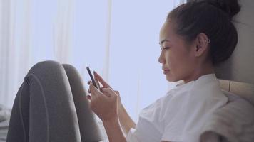 ung solbränna asiatisk kvinna sitta ner på knäna med smartphone på sängen, nätverksanslutning online, nya flöden upptagna morgonmeddelanden, bekvämt sovrumsfönster dagsljus, ensamt sovrumsstämning video
