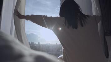 asiatisk kvinna vaknar och öppnar fönstergardiner sträcker armarna på sängen på morgonen, titta ut genom fönstret med utsikt över stadsgatan, höghus lägenhet, börja dagen med energi video