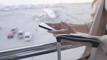 close up business woman hands use mobile phone texting chatting while waiting for the flight, standing inside airport terminal business trip, cellphone and travel luggage and airplane on background video