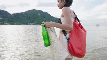 Asiatisk ung kvinna som samlar in plastpåse och glasflaska från stranden på ön vid havet medan hon reser och plockar upp skräp frivilligt återvinning av sopor. ekologisk miljömedvetenhet