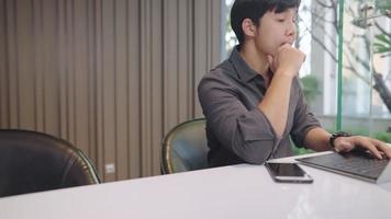 nachdenklicher, uninspirierter asiatischer geschäftsmann, der an eine neue idee zur lösung von arbeitsproblemen denkt, konzentriertes denken vor dem laptop, junger unternehmer in formeller kleidung, der allein am schreibtisch sitzt video