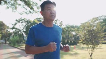 Asian senior man running inside the running track at the park on a sunny day in slow motion. Retirement lifestyle activity. Health care motivation concept,  endurance cardio exercise, front view video