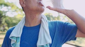 nahaufnahme senior asiatischer mann trägt blaues t-shirt und trinkt frisches kaltes wasser aus plastikflasche während des trainings im freien im park, älteres training, erfrischendes getränk, rehydrierung des lebensstils im ruhestand