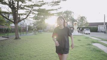 mujer asiática corriendo en el parque de la calle del vecindario sobre hierba verde bajo las sombras de los árboles, al atardecer, ejercitando la motivación, perdiendo peso, ejercitando el acondicionamiento corporal, controlando el peso de la dieta, cámara lenta video