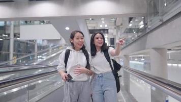 Asian attractive teenage tourist friends in holiday trip looking pointing finger forward inside modern airport hall. Lost beautiful travelers with baggage on escalator finding direction to checking video