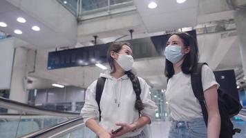 Asian beautiful friends wear medical mask happily talking while using escalator walkway, female backpackers cheerfully traveling together, vacation going abroad, teenager girls on exploring the world, video