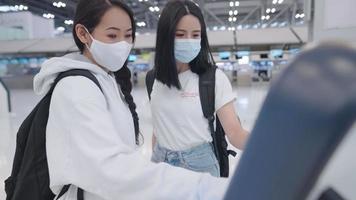 Two young asian female friends arrived at airport terminal, using automatic kiosk machine for checking in, receive flight ticket boarding pass, modern technology and transportation, new normal travel