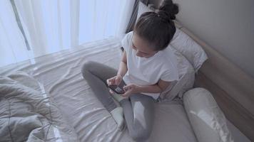 giovane donna asiatica con i capelli legati seduti in camera da letto, uso del cellulare per collegarsi online, connessione di rete, risposta commenti nuovi feed, messaggistica mattutina impegnata, distanza sociale isolata e sola video