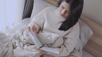 Young adult Asian woman reading book sitting on the bed, wearing warm sweater, messy blanket sheets pillow on the back, morning spending free time on the weekend, comfortable bedroom window day light video
