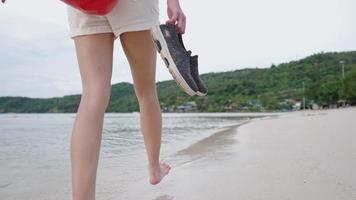 shot van een jong meisje met schoenen die op blote voeten lopen op het tropische zandstrand, zomerweer in Azië, waterdichte tas voor extreem reizen, ontspannende wandeling langs de kustkust, eilandretraite video