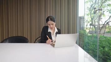 aantrekkelijke aziatische ondernemersvrouw die een marketingonderzoek doet terwijl ze alleen zit in een moderne kantoorkamer, makelaar regelt volgens klantenschema, reageert op het schrijven van een e-mail video